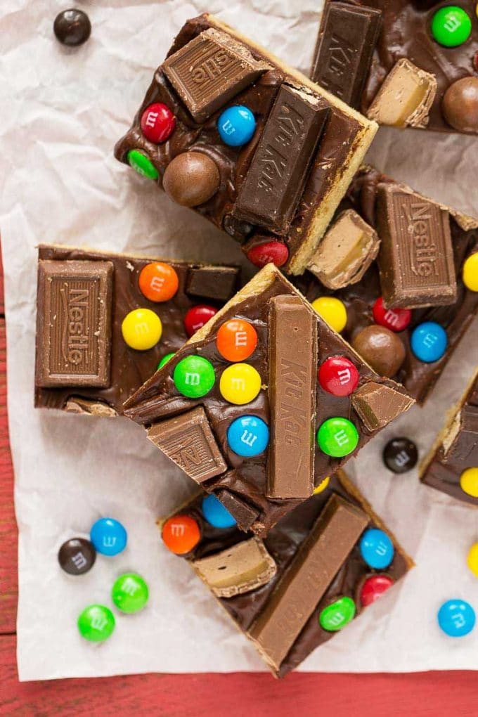 Several candy bar blondies stacked on a paper topped with rich chocolate frosting and a variety of candies.