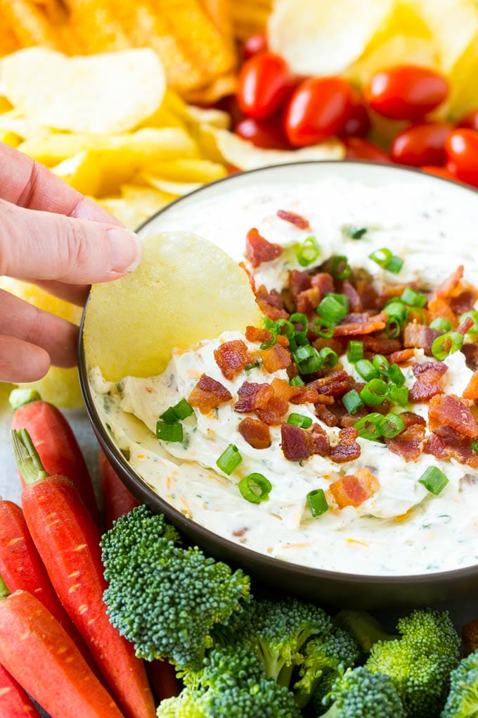 Bacon Ranch Dip Recipe | Homemade Ranch Dip | Easy Dip Recipe | Cheddar Ranch Dip #ranch #ranchdip #bacondip #dip #snack #edsug #baconranchdip A hand scooping up bacon ranch dip with a potato chip.