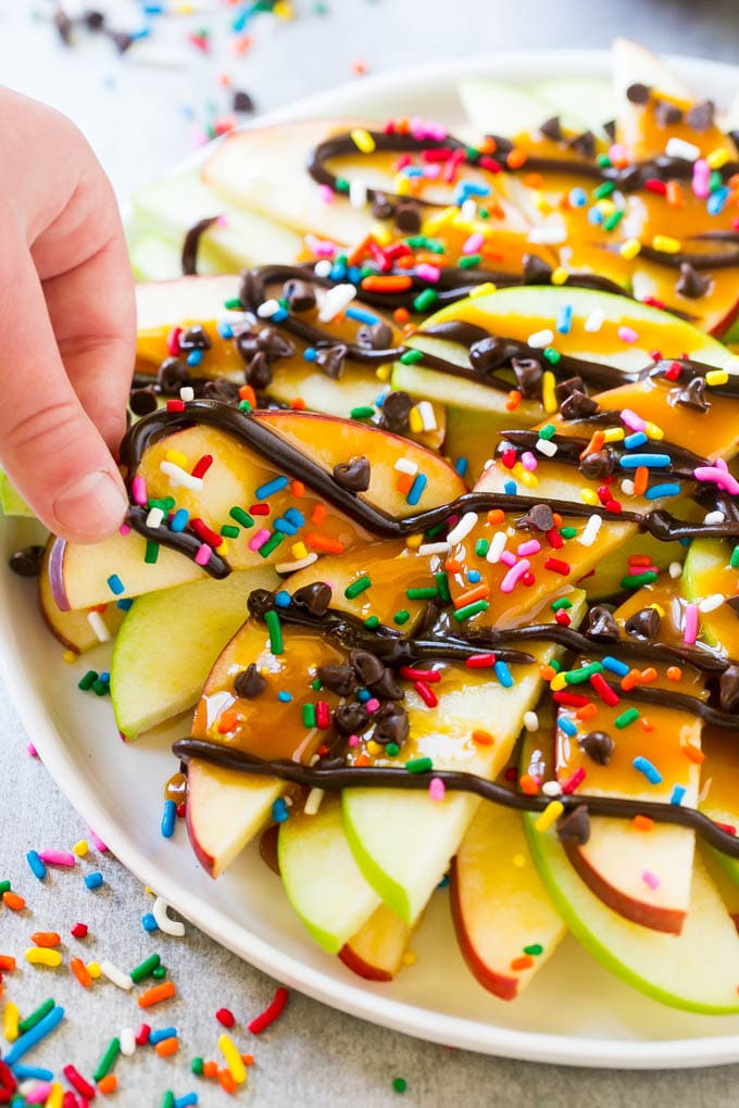 Apple Nachos Recipe | After School Snack | Caramel Apples #apple #apples #caramelapple #snack #dessert #edsug A plate of caramel apple nachos with a hand reaching out to take one.
