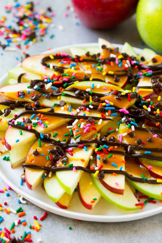 Apple Nachos Recipe | After School Snack | Caramel Apples #apple #apples #caramelapple #snack #dessert #edsug A plate of red and green sliced apples drizzled with caramel and chocolate, then topped with rainbow sprinkles and chocolate chips.