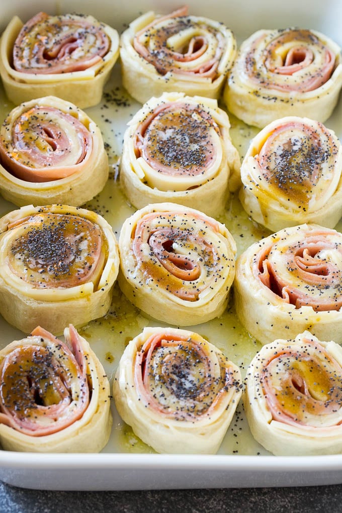 Ham and Cheese Pinwheels Recipe | Ham and Cheese Roll Ups | Ham and Cheese Rolls | Ham and Cheese Bread Rolls #ham #cheese #appetizer #pinwheels #edsug Dough rolled up with ham and cheese and topped with a poppy seed butter in a ceramic dish.