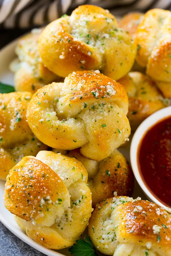 Garlic Knots Recipe | Garlic Rolls | Garlic Butter Rolls | Garlic Bread Recipe #garlic #garlicbread #garlicknots #breadsticks #edsug A plate of garlic knots slathered in butter next to a cup of marinara sauce.