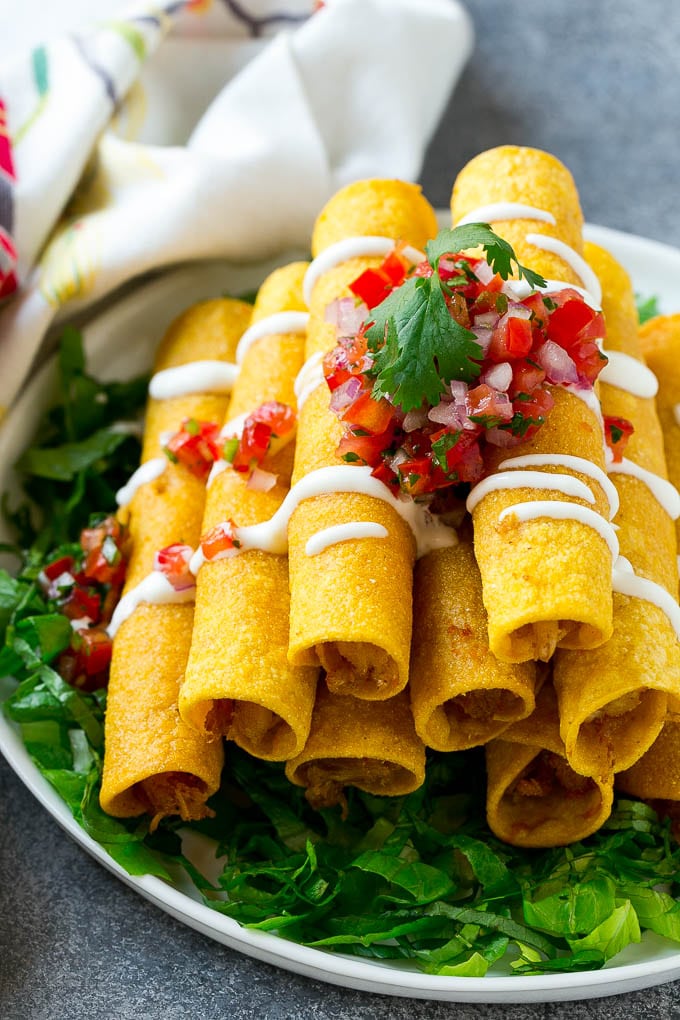 Chicken Taquitos Recipe | Baked Chicken Taquitos | Fried Chicken Taquitos | Mexican Appetizer | Chicken Tortillas #taquitos #chicken #appetizer #tortillas #mexicanfood #edsug A stack of chicken taquitos with a drizzle of sour cream and pico de gallo topping.