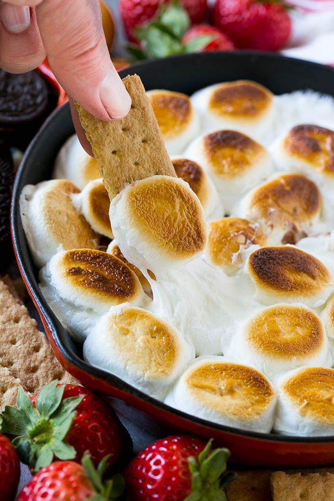 S'mores Dip Recipe | S'mores Skillet | Baked S'mores #smores #dip #chocolate #marshmallow #dessert #edsug A graham cracker topped with melted marshmallow dipping into a skillet of s'mores dip.