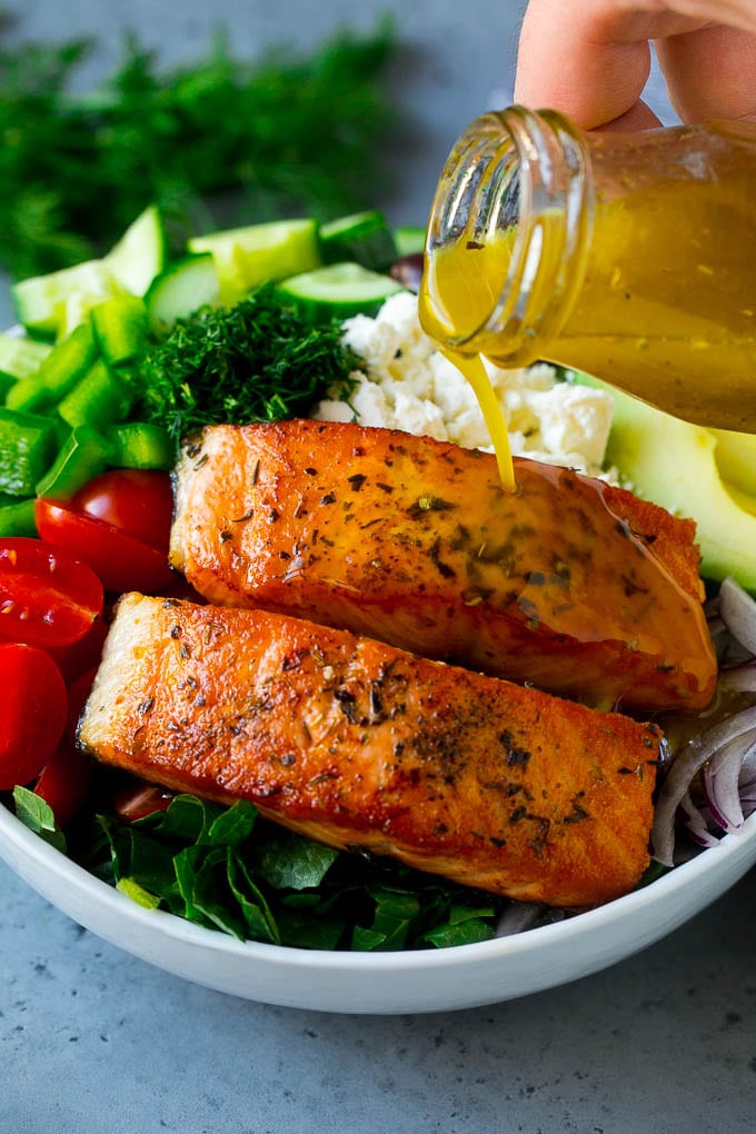 Greek Salmon Salad Recipe | Low Carb Salmon Recipe | Salmon Salad #salmon #seafood #fish #lowcarb #keto #lunch #edsug #healthy Salmon fillets on top of greens with dressing being poured over the top.