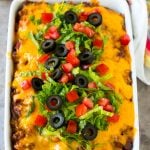 Taco casserole in a baking dish topped with melted cheese, lettuce, tomato and olives.