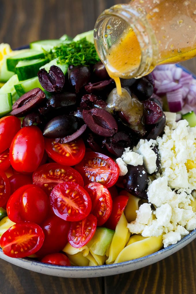 Greek Tortellini Salad Recipe | Pasta Salad | Tortellini Salad | Greek Salad #greek #tortellini #pasta #salad #cucumbers #olives #dinner #edsug Salad dressing being poured over a bowl of tortellini and vegetables.