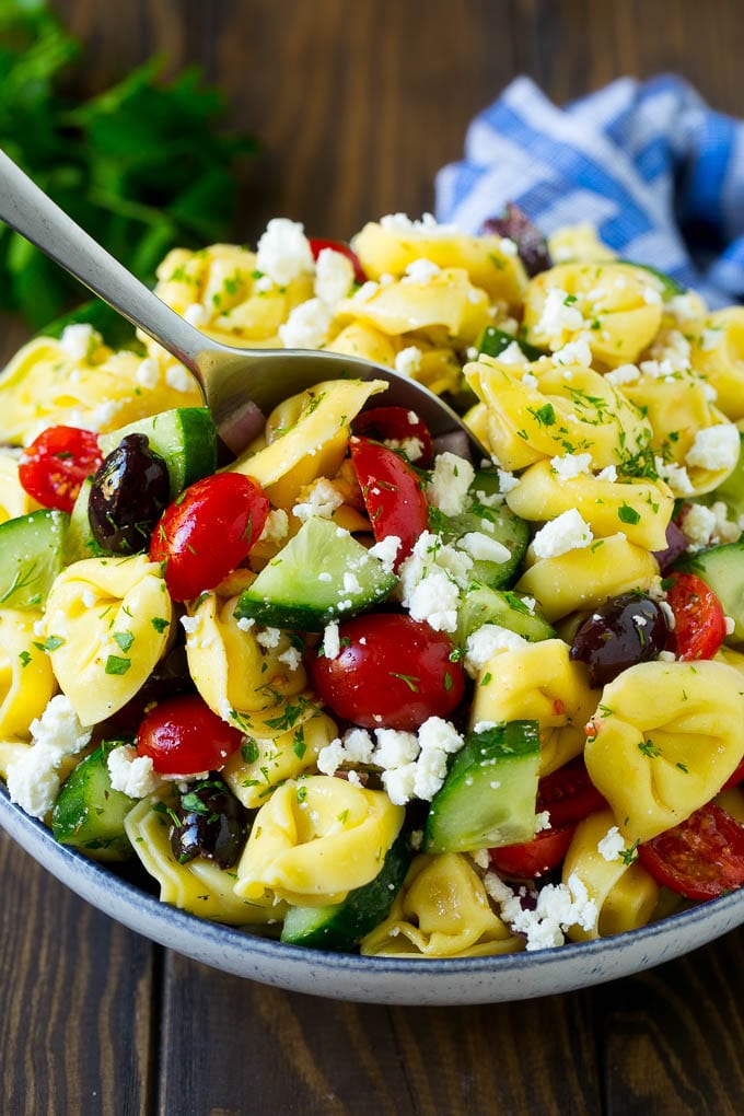 Greek Tortellini Salad Recipe | Pasta Salad | Tortellini Salad | Greek Salad #greek #tortellini #pasta #salad #cucumbers #olives #dinner #edsug A bowl of Greek tortellini salad with a serving spoon in it.