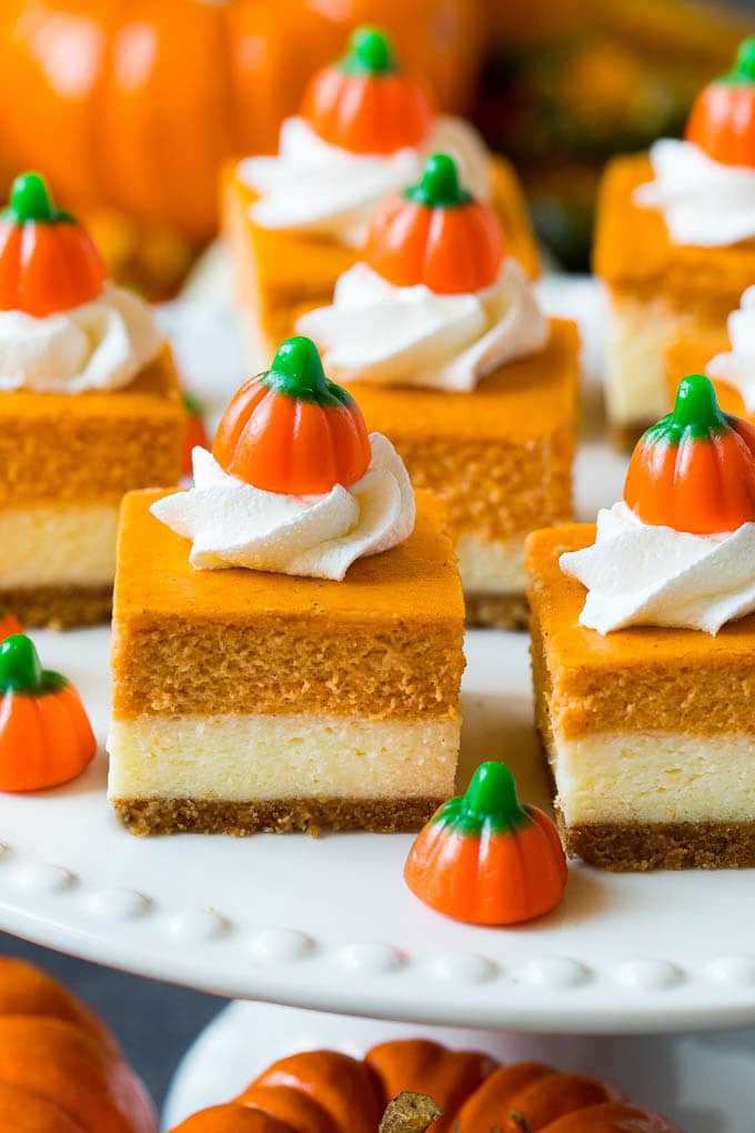These pumpkin cheesecake bars are layers of graham cracker crust, vanilla cheesecake and pumpkin cheesecake, all baked together to create an impressive dessert. The perfect alternative to pumpkin pie on Thanksgiving day! A platter of pumpkin cheesecake bars topped with whipped cream and candy pumpkins.
