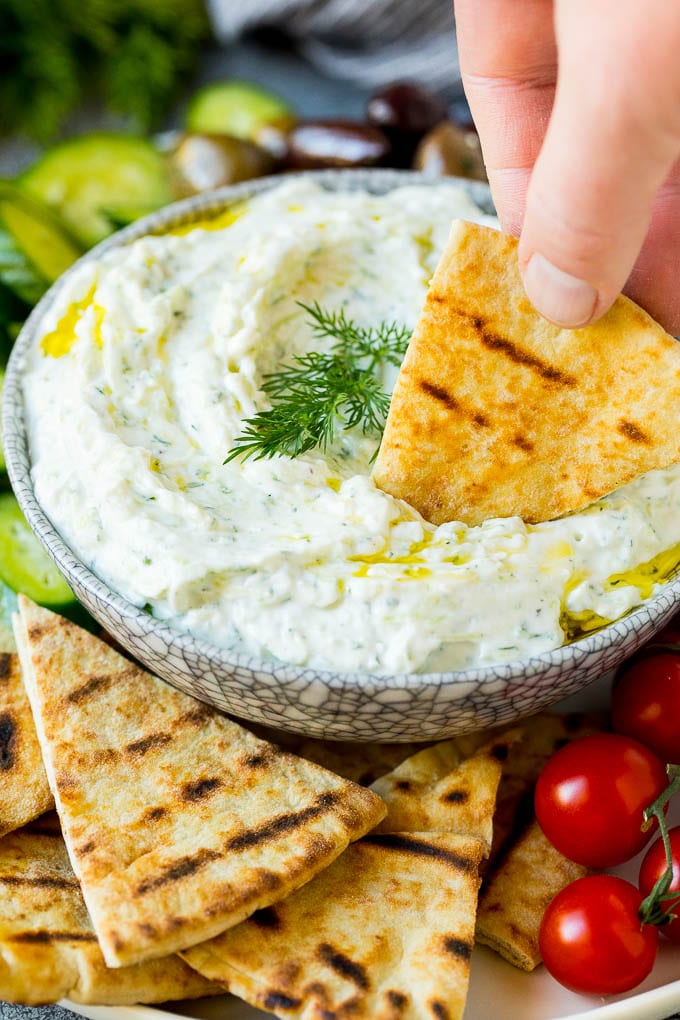 The best homemade tzatziki sauce with cucumber and herbs mixed into Greek yogurt. #appetizer #edsug A hand dipping pita bread into a bowl of tzatziki.