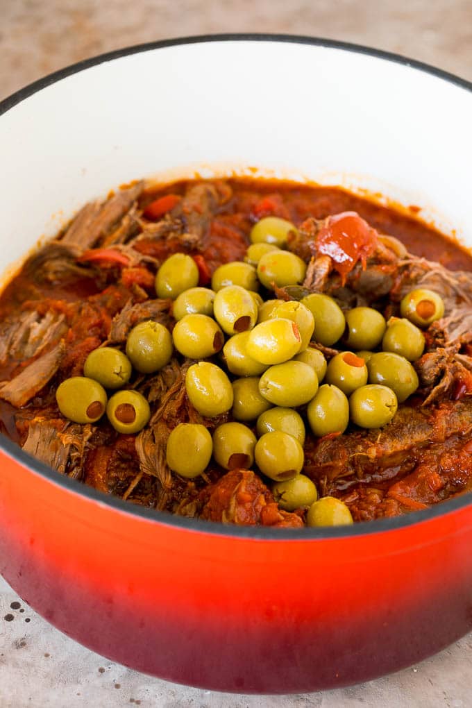 This ropa vieja recipe is a traditional dish of beef roast that's simmered with tomatoes and seasonings until tender, then shredded and served with olives. A pot of shredded beef with whole green olives on top.