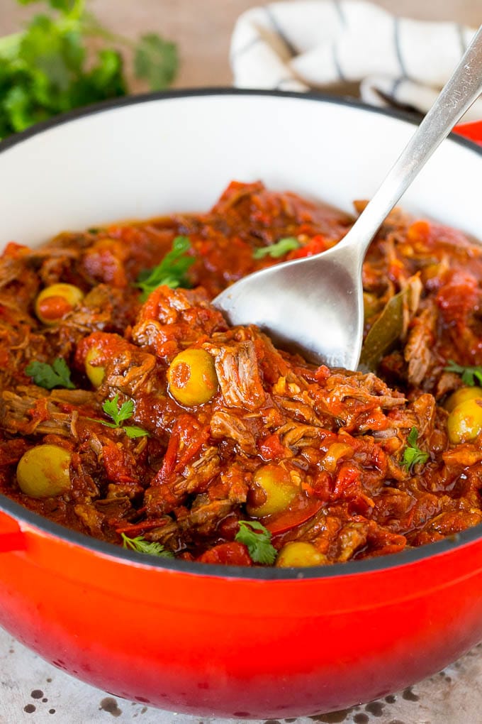 This ropa vieja recipe is a traditional dish of beef roast that's simmered with tomatoes and seasonings until tender, then shredded and served with olives. A serving spoon in a pot of ropa vieja with a garnish of cilantro.