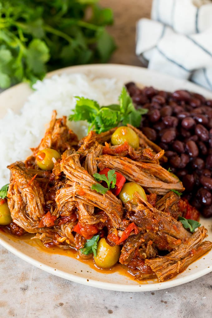 This ropa vieja recipe is a traditional dish of beef roast that's simmered with tomatoes and seasonings until tender, then shredded and served with olives. A plate of ropa vieja served with rice and black beans.