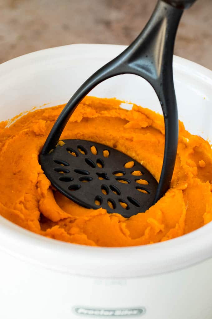 This crock pot sweet potato casserole is yams mashed with sweetener, vanilla and spices, then finished off with a brown sugar pecan topping. A potato masher that is mashing sweet potatoes in a crock pot.