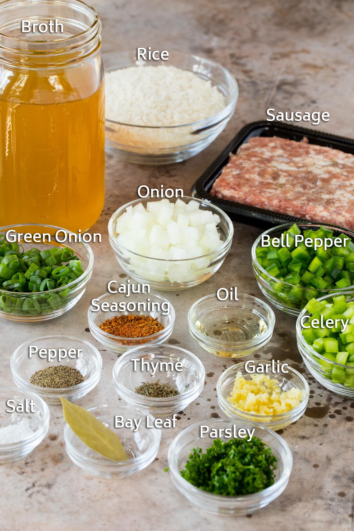 Bowls of ingredients including vegetables and seasonings and rice, as well as a container of sausage and jar of broth.