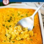 A dish of cornbread casserole and a metal spoon scoop some of the casserole.