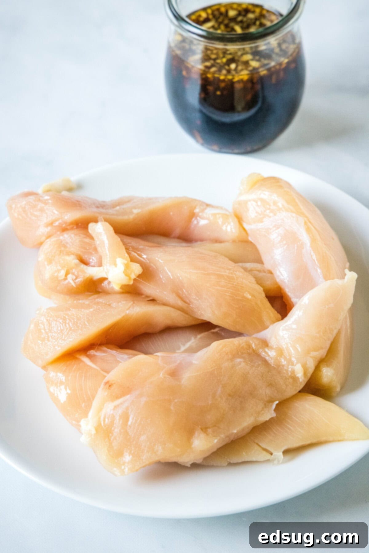 Chicken pieces on a white plate with a jar of teriyaki sauce in the background, ready for marinating.
