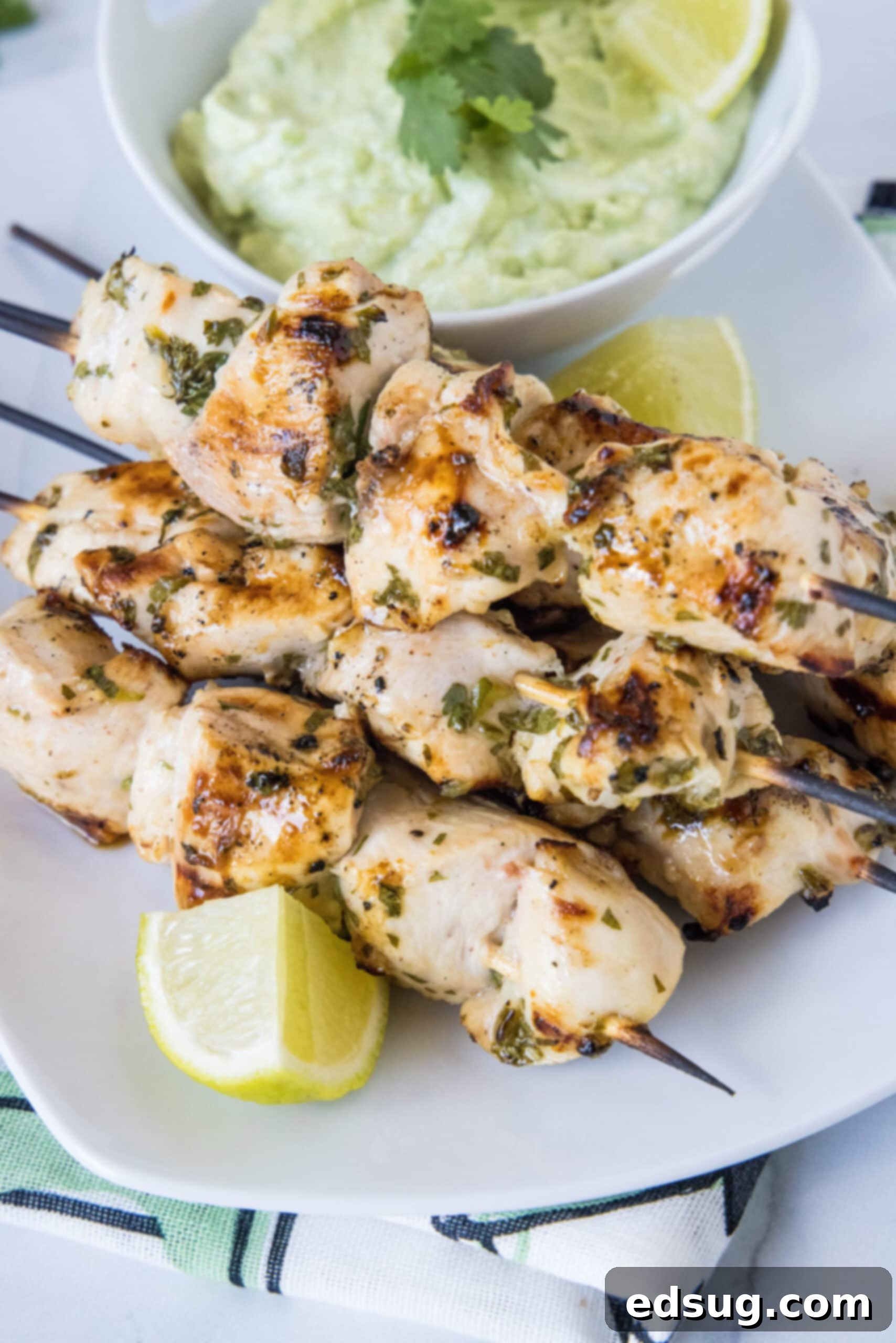A plate of grilled cilantro lime chicken skewers next to a lime wedge, with a bowl of avocado dipping sauce in the background.