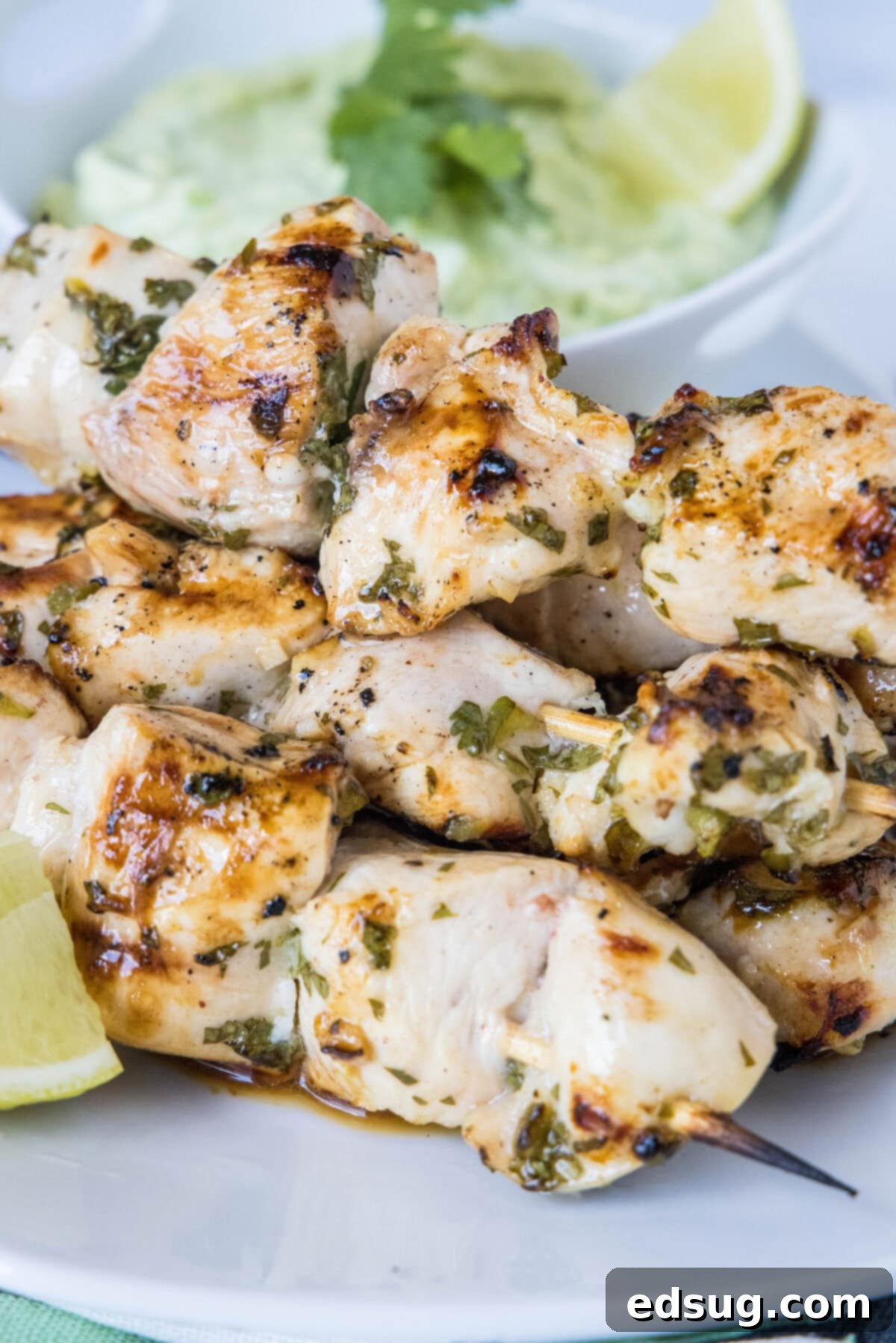 A plate of grilled cilantro lime chicken skewers next to a lime wedge, with a bowl of avocado dipping sauce in the background.