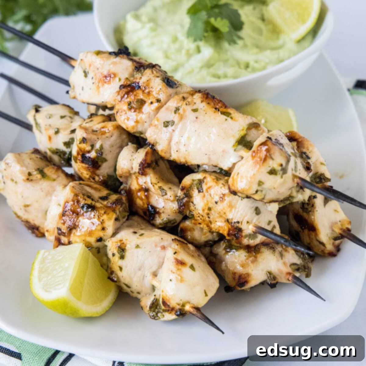 A plate of grilled cilantro lime chicken skewers next to a lime wedge, with a bowl of avocado dipping sauce in the background.