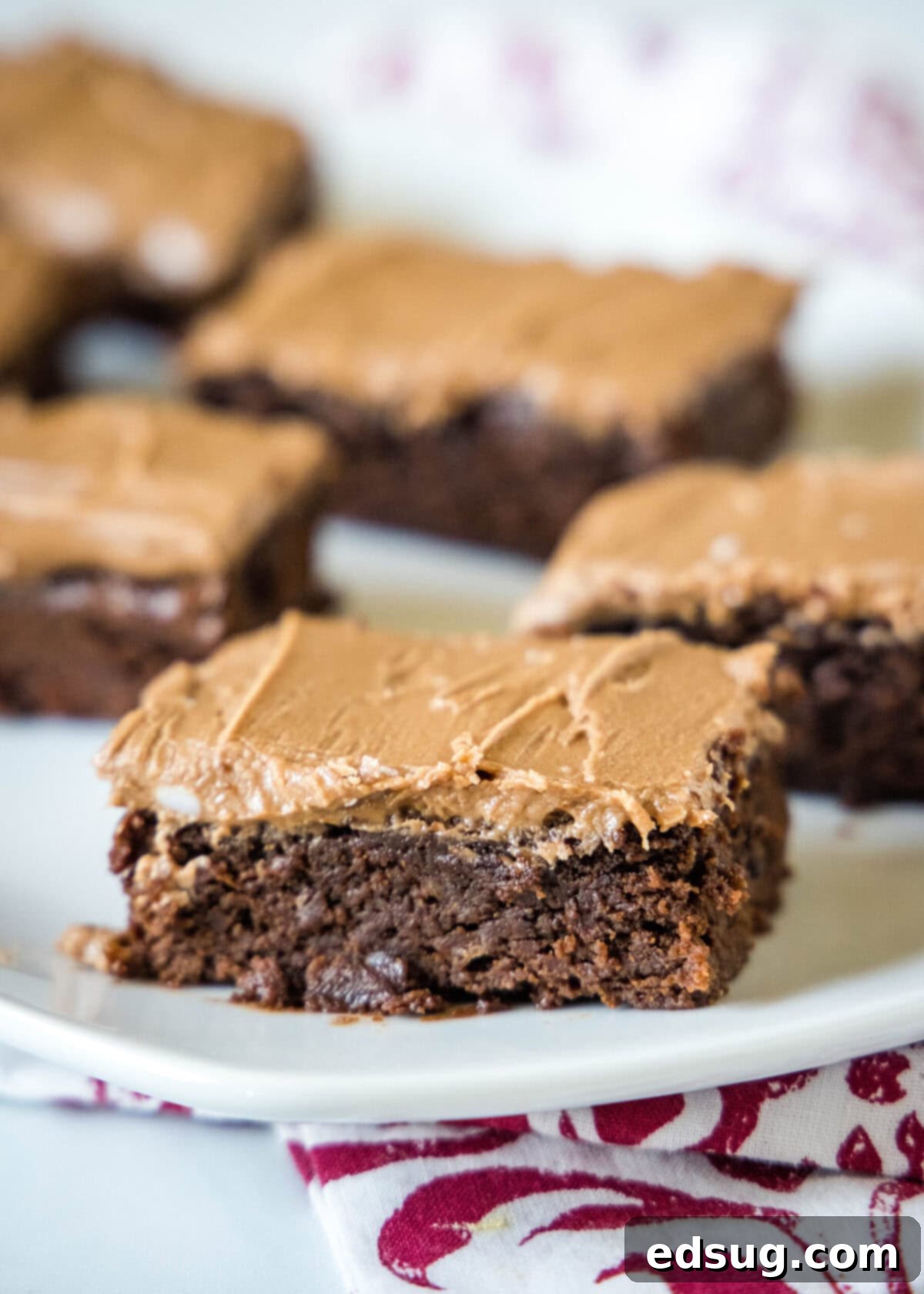 Close up of a single brownie square with chocolate frosting, with more brownie squares blurred in the background.