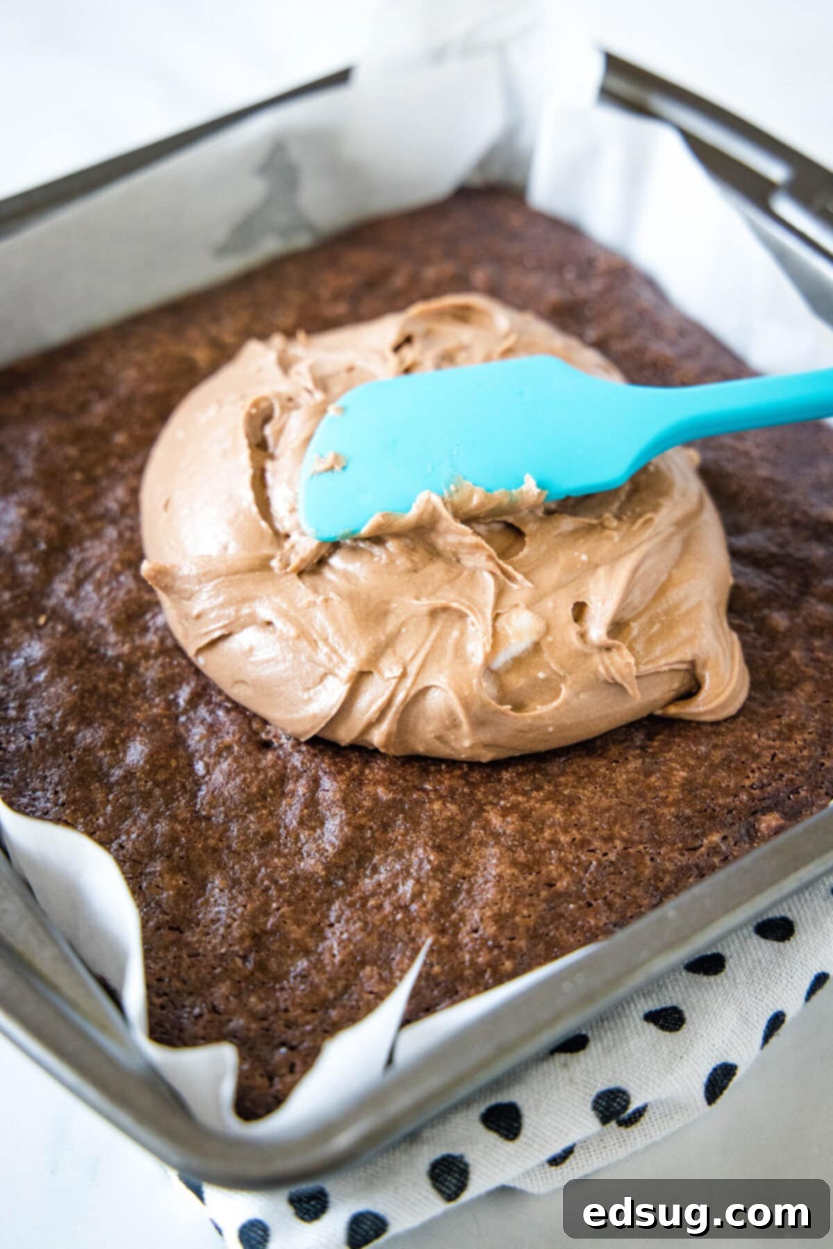 A rubber spatula spreading rich chocolate frosting over a pan of warm brownies.