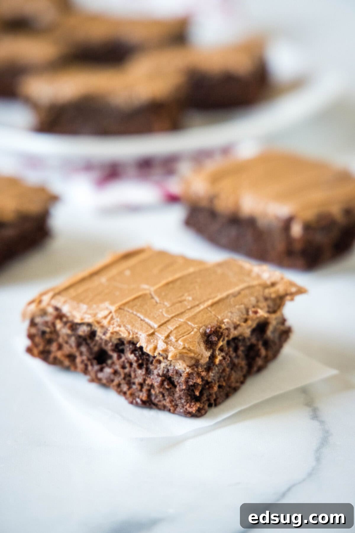 A single dairy-free brownie square with chocolate frosting, with a plate of more brownies in the blurred background.
