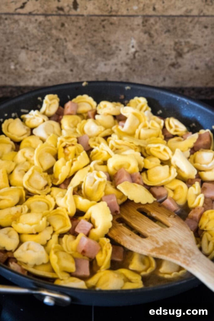 A wooden spatula stirring together cheese tortellini, diced ham, and onions in a skillet.