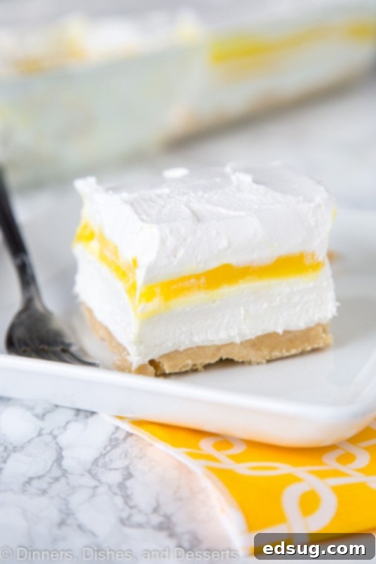 A slice of creamy lemon lasagna dessert on a white plate, showcasing distinct layers of golden crust, white cream cheese, yellow lemon pudding, and a light whipped topping.