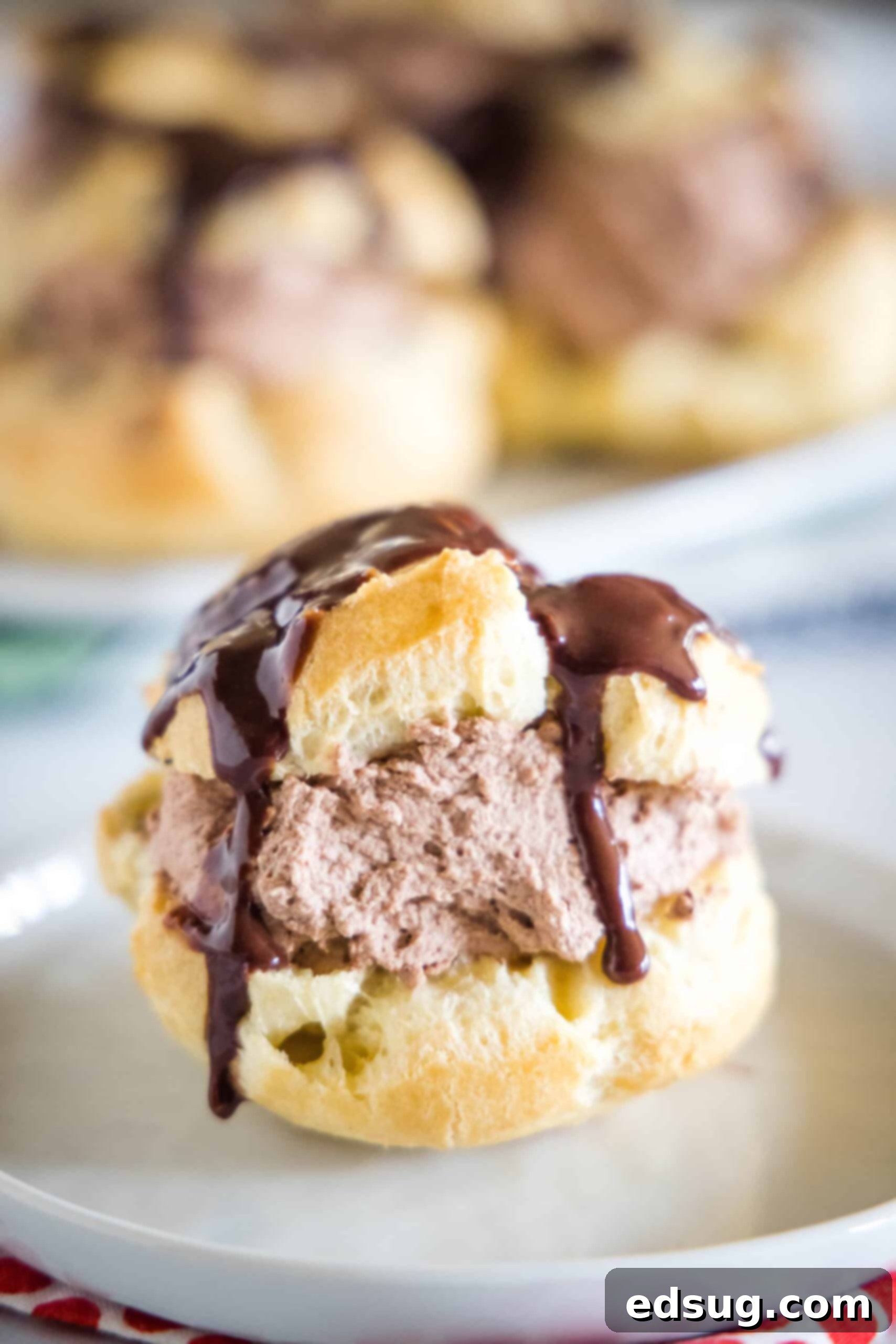 A chocolate cream puff topped with chocolate ganache on a white plate, with more cream puffs on a plate in the background.