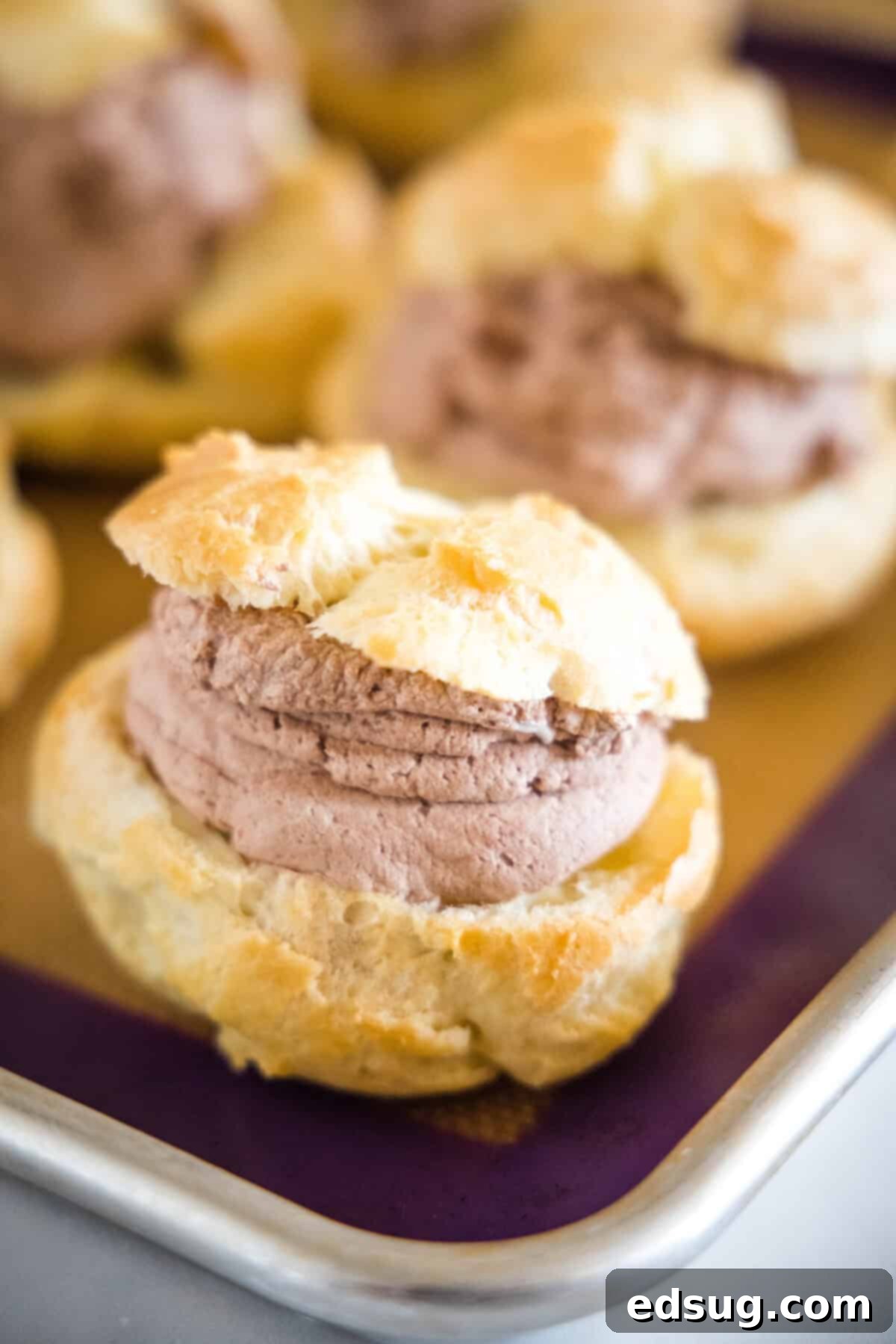 Close up of a filled chocolate cream puff in the corner of a baking pan with more filled puffs in the background.
