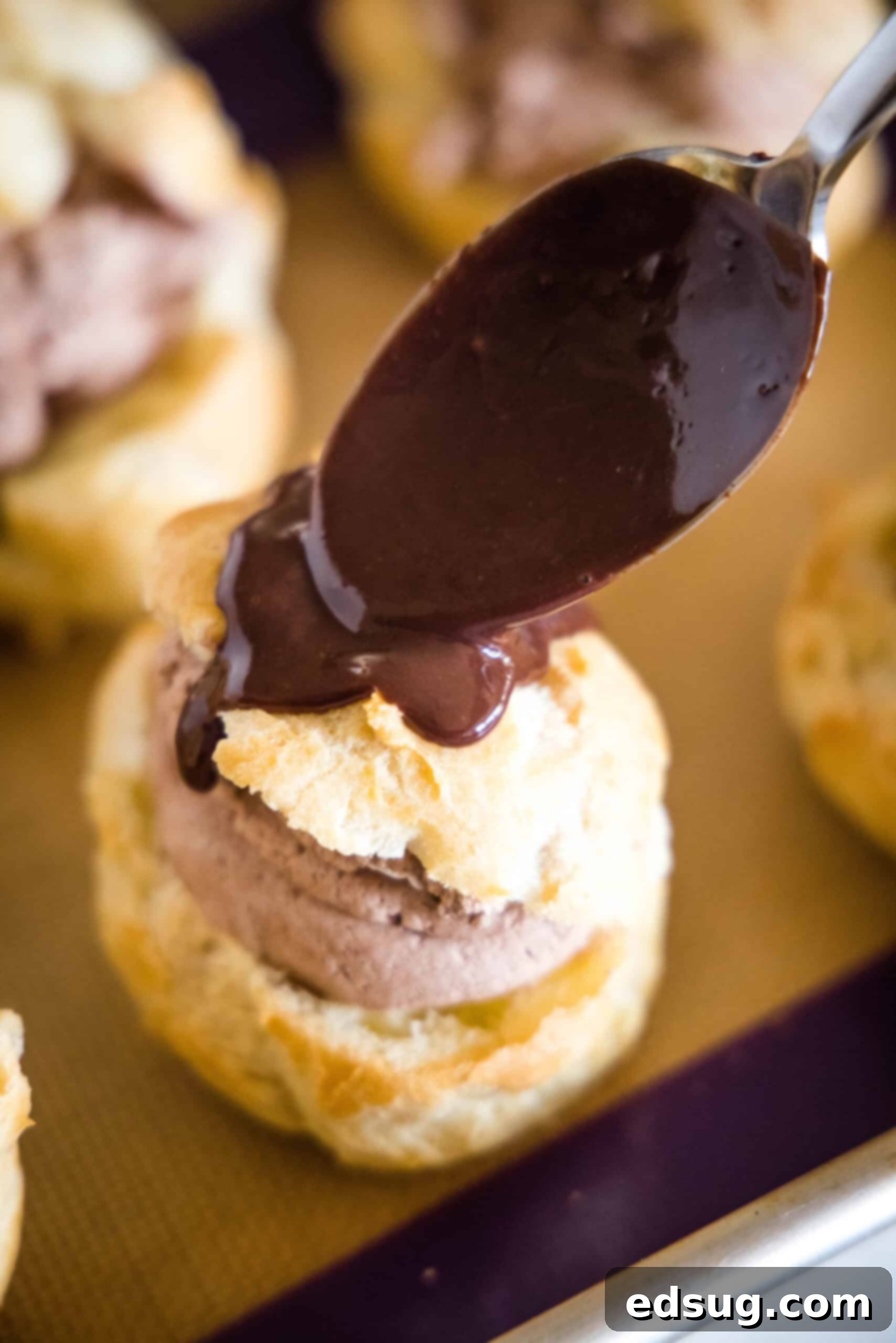 A spoon dripping chocolate ganache on top of a prepared chocolate cream puff.