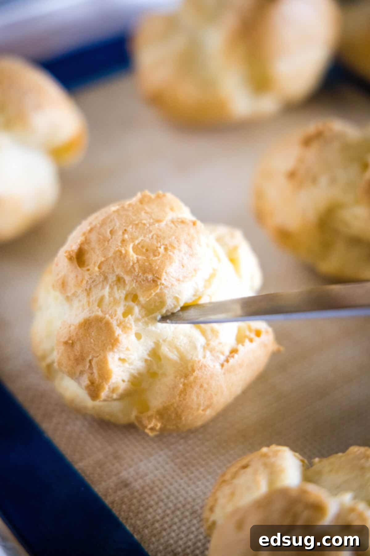 A knife piercing the side of a baked cream puff on a baking sheet.