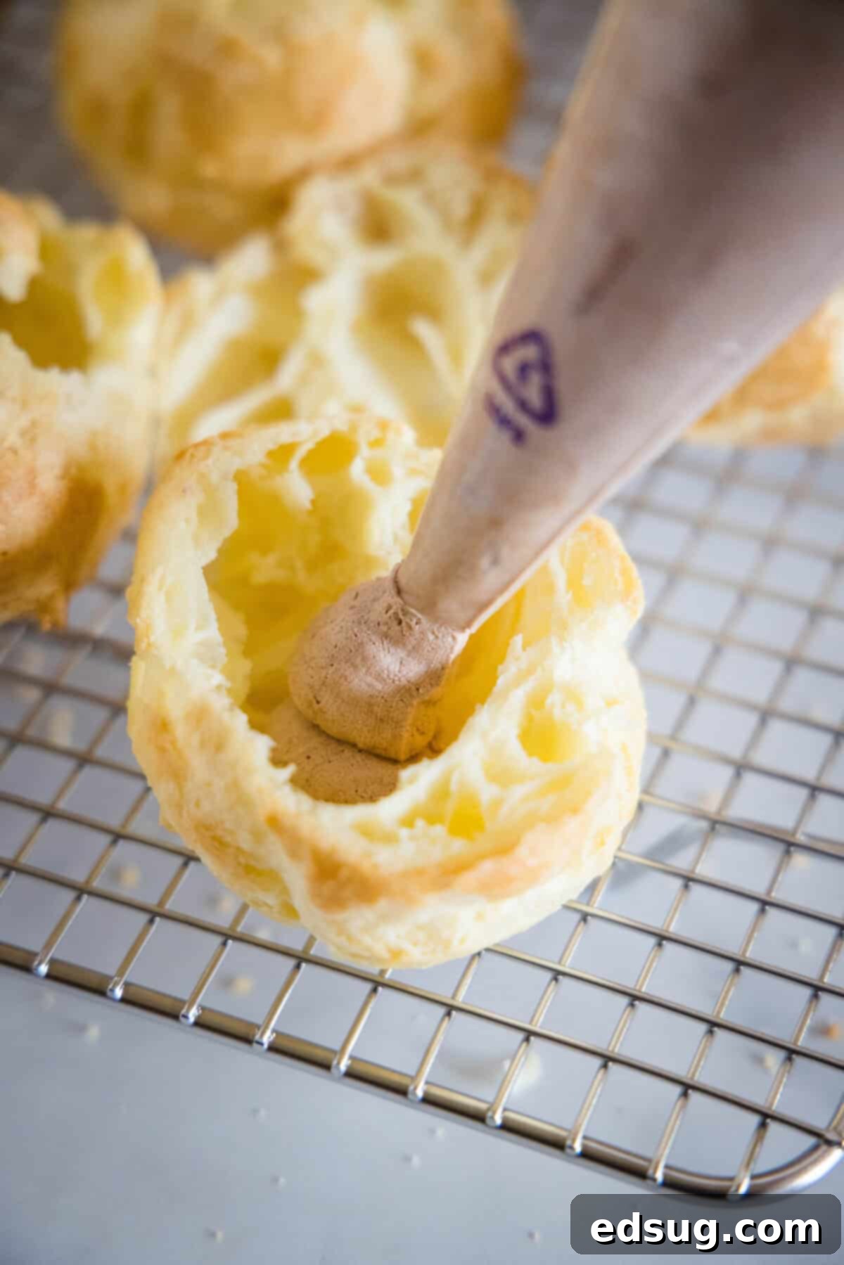 The tip of a piping bag filled with chocolate whipped cream held over an open cream puff on a wire rack.