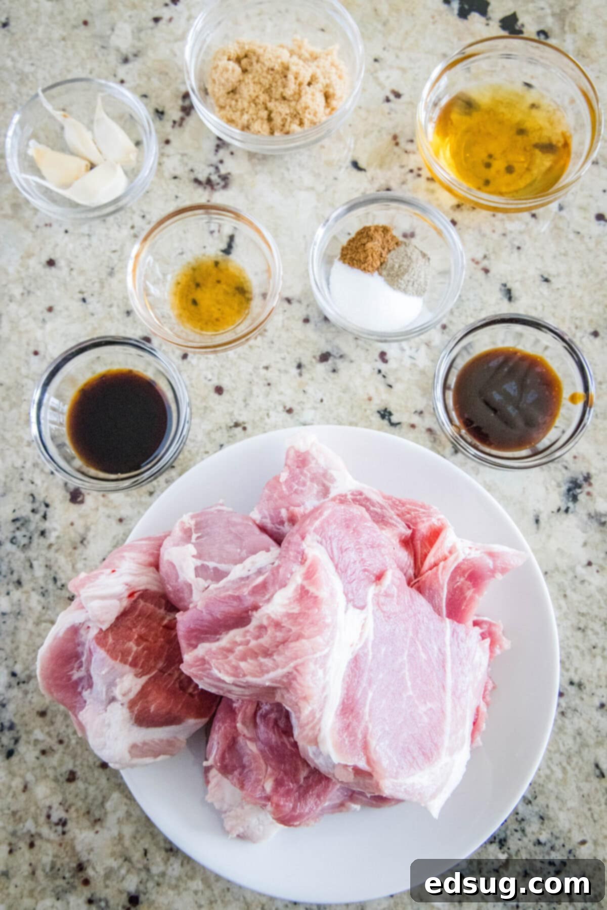 This easy char siu pork recipe is tender, juicy, and loaded with your favorite Chinese flavors like hoisin and five-spice powder. Overhead view of the ingredients needed for char siu pork: a plate of raw pork shoulder, a bowl of soy sauce, a bowl of hoisin, a bowl of garlic, a bowl of sesame oil, a bowl of brown sugar, a bowl of honey, and a bowl with salt, pepper, and five-spice.