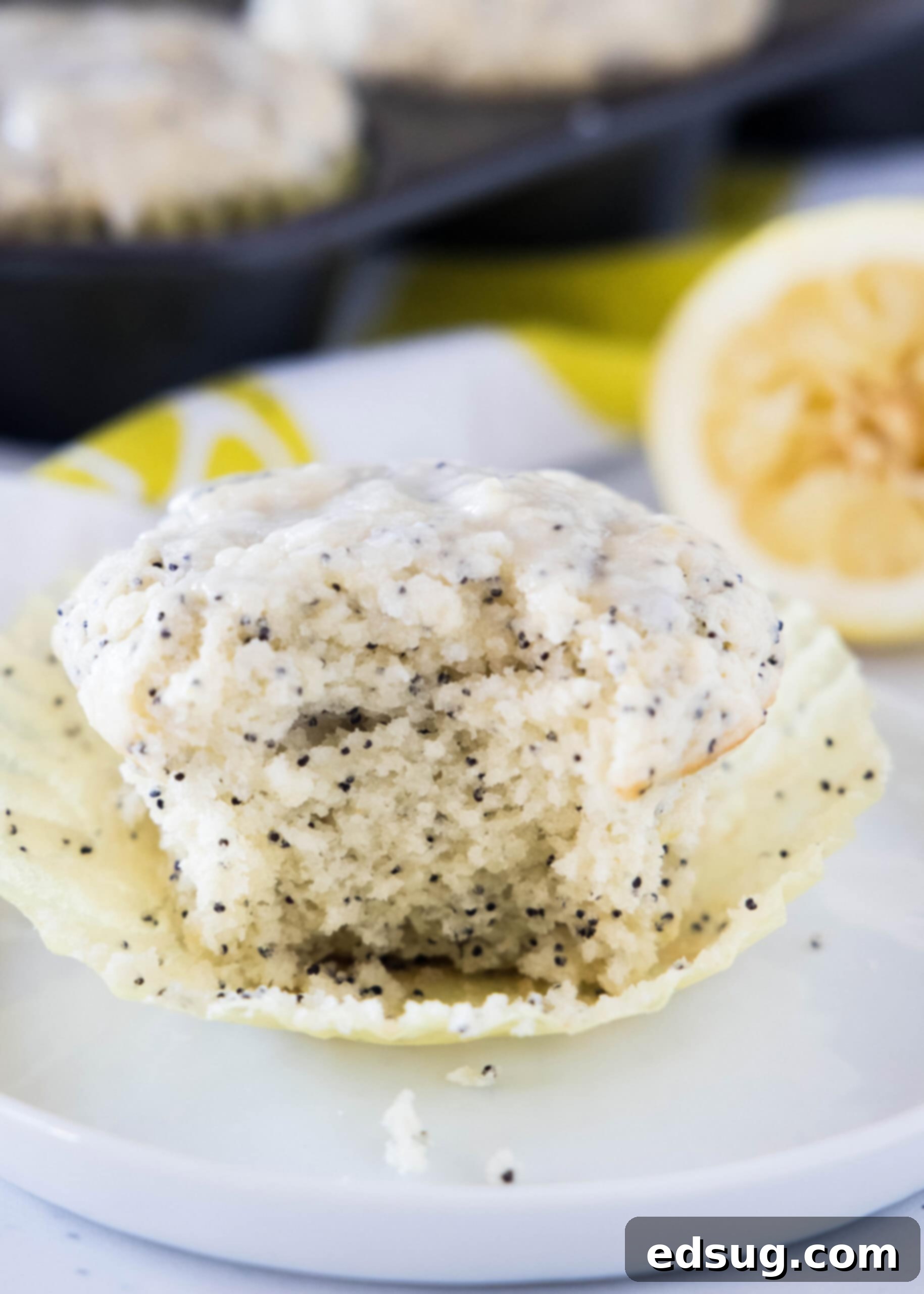 Close up of an unwrapped lemon poppy seed muffin on a white plate, with a bite missing. The muffin is golden brown with a visible lemon glaze and poppy seeds.