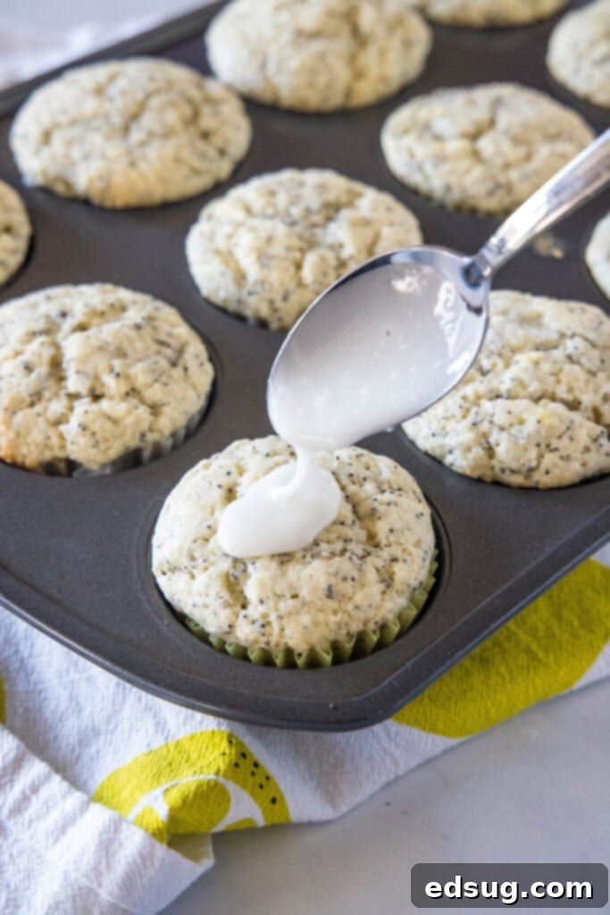 A spoon expertly drizzling a glistening lemon glaze over a warm lemon poppy seed muffin in a muffin tin.