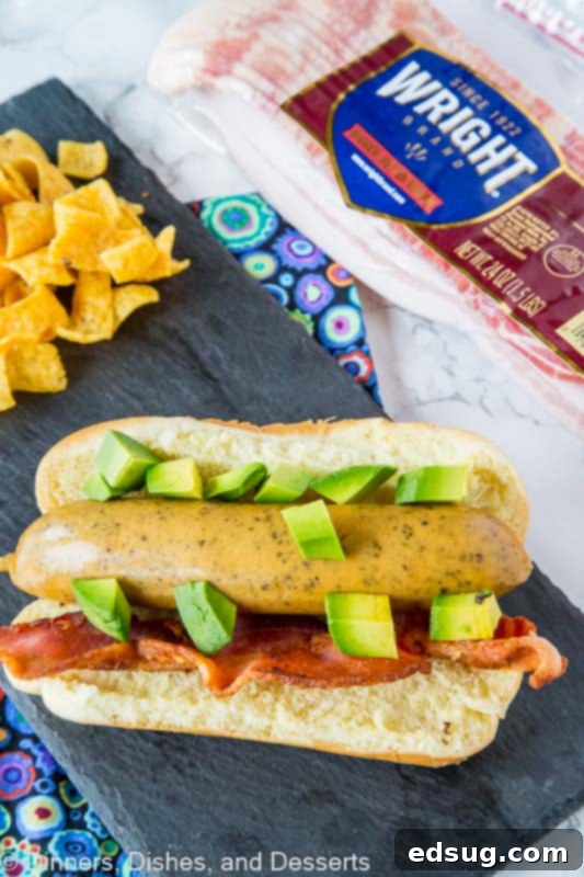 A single Bacon Avocado Beer Brat on a cutting board, garnished with fresh avocado and bacon bits.