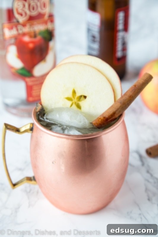 A copper mug of Apple Cider Moscow Mule with apple slices and cinnamon stick next to fresh apples and cider