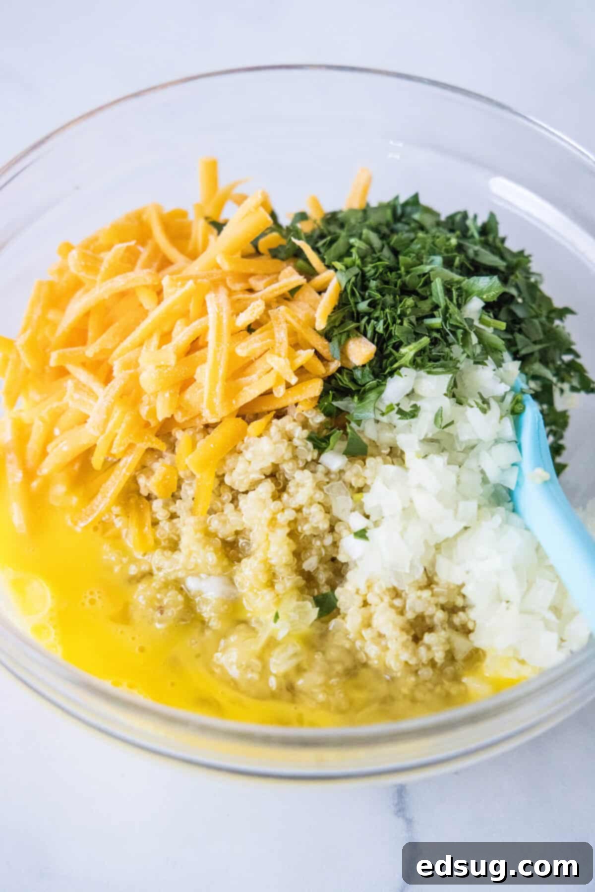 All the cooked and prepared ingredients for quinoa bites, including quinoa, cheese, herbs, and egg, thoroughly mixed in a large glass bowl.