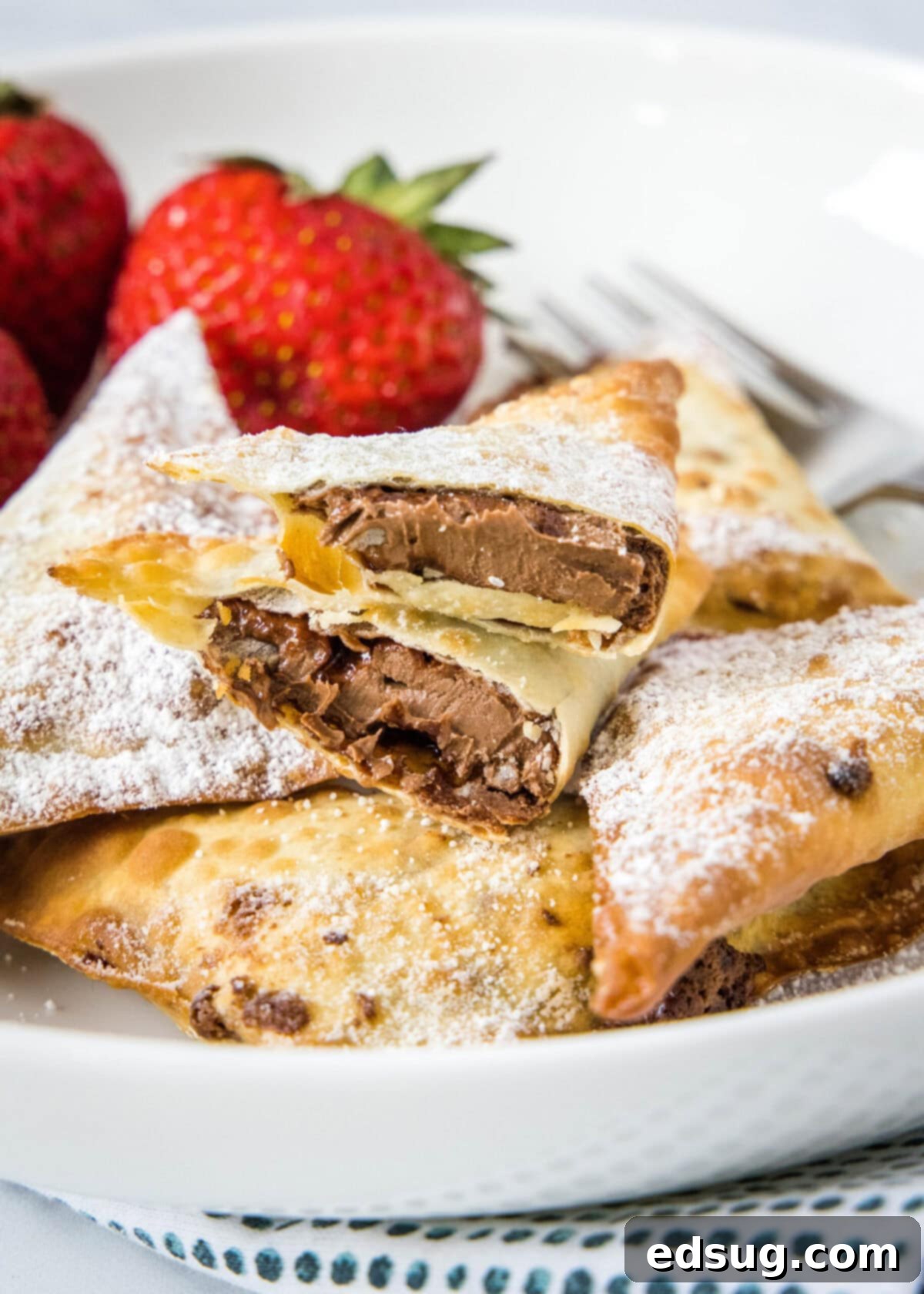 A plate with a pile of wontons covered in powdered sugar, with one cut in half and full of Nutella, with strawberries in the background, showcasing the crispy exterior and gooey chocolate-hazelnut filling.