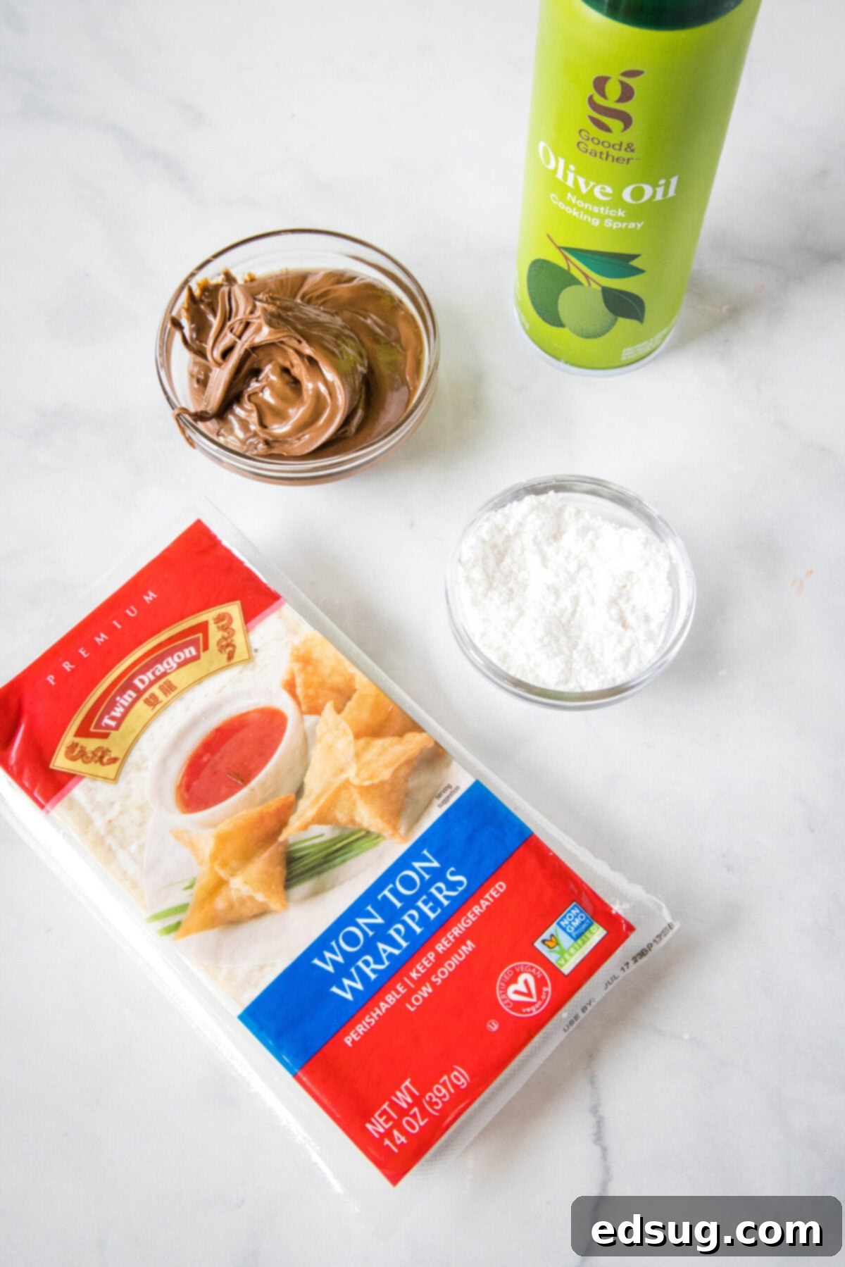 Overhead view of the simple ingredients needed for delicious dessert wontons: a package of wonton wrappers, a bowl of creamy Nutella, a bowl of fine powdered sugar for dusting, and a bottle of olive oil spray for crisping.