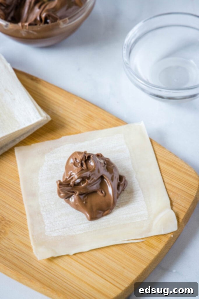 A mound of Nutella precisely placed in the middle of a wonton wrapper, next to a stack of unused wrappers, a bowl of Nutella, and a small bowl of water for sealing, illustrating the initial filling step.