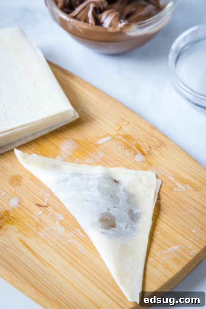 An uncooked wonton wrapper perfectly folded into a triangle, filled with Nutella and sealed, placed next to a stack of fresh wrappers and a bowl of Nutella, demonstrating the folded stage before air frying.