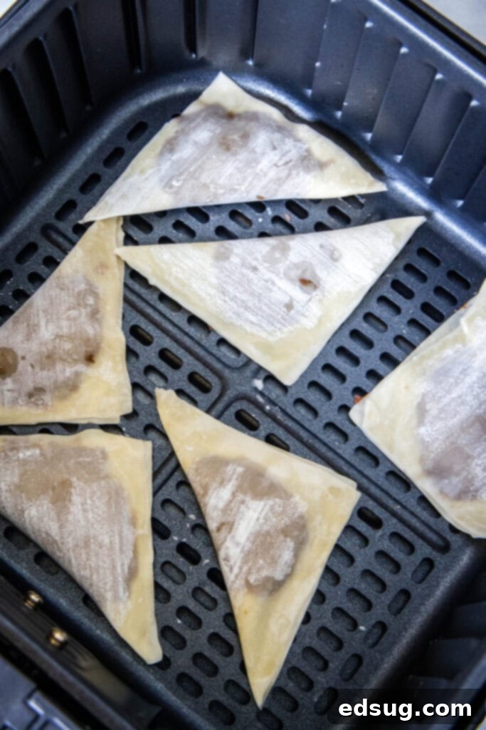 Six uncooked wonton wrappers, filled with Nutella and neatly folded, arranged in a single layer within an air fryer basket, ready for cooking to ensure even crisping.