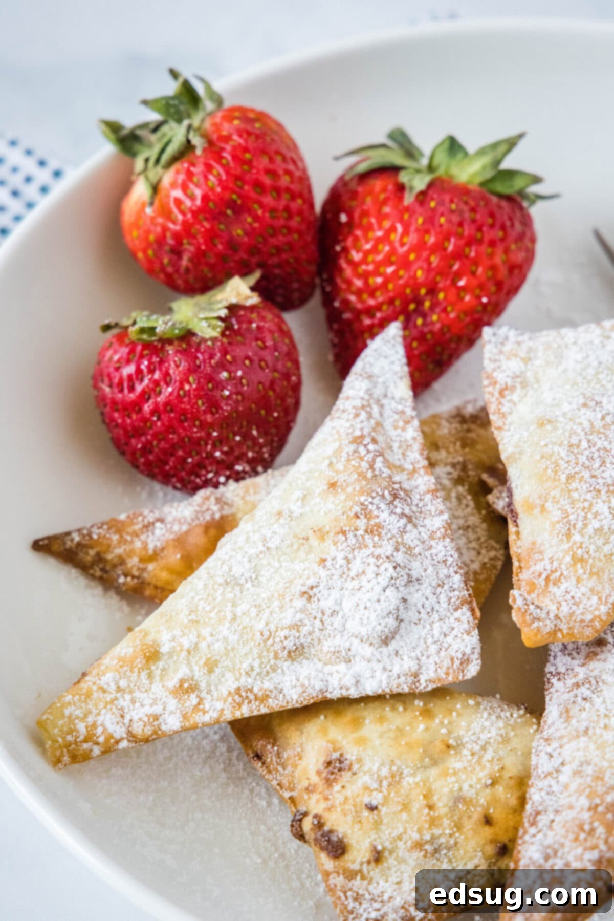 Overhead view of a few golden, powdered-sugar-dusted Nutella wontons, beautifully arranged next to three fresh, ripe strawberries, highlighting a perfect serving suggestion.
