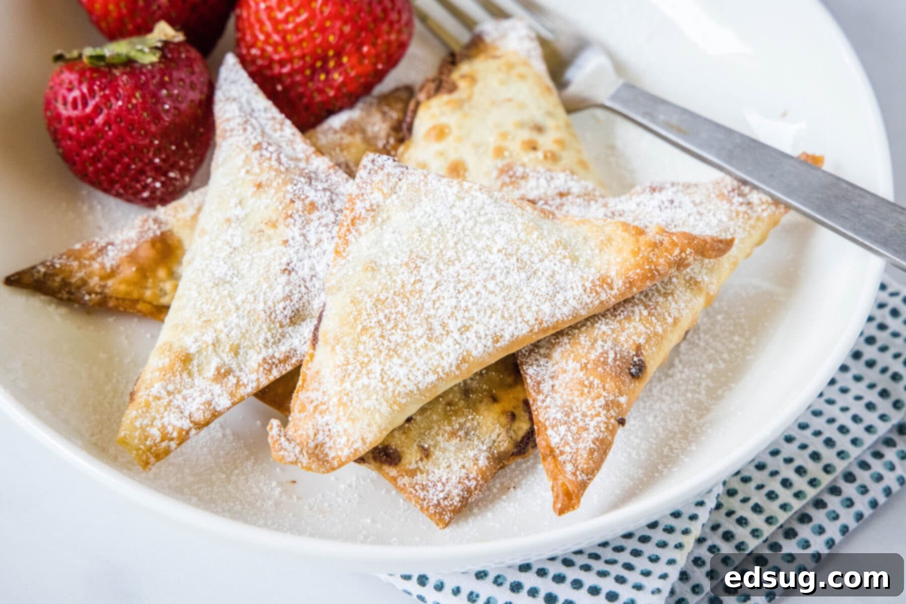 A beautifully presented plate of golden dessert wontons, generously covered in powdered sugar, with fresh strawberries and a fork beside them, ready to be enjoyed.