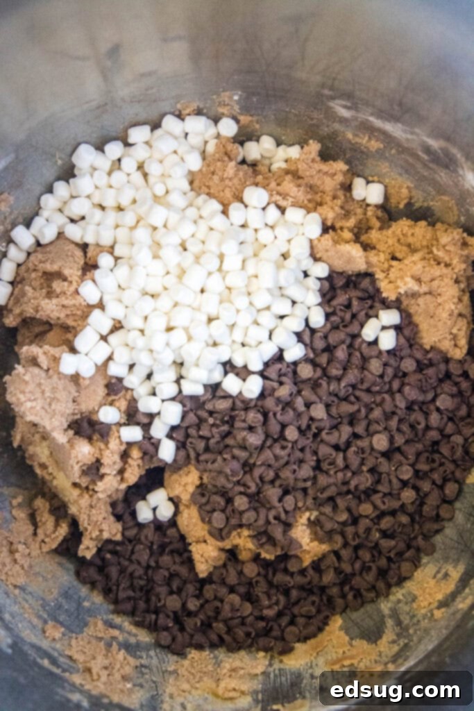 Decadent Hot Chocolate Cookies 3 Hands adding a generous amount of chocolate chips and mini marshmallow bits to a bowl of hot chocolate cookie dough, ready for mixing.