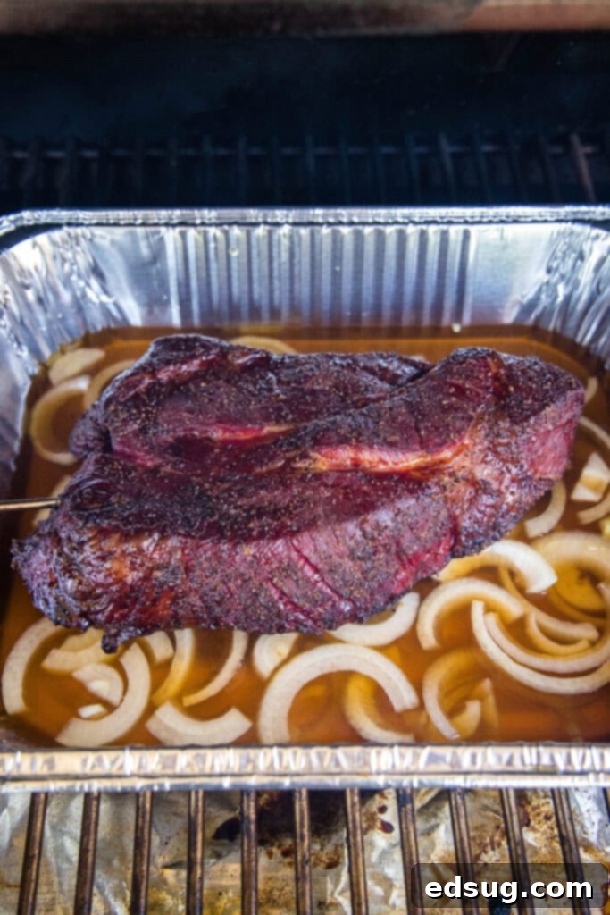 Chuck roast in an aluminum pan with sliced onions and beef broth, ready for the smoker.