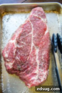 A raw chuck roast evenly coated in a Texas-style dry rub on a baking tray.